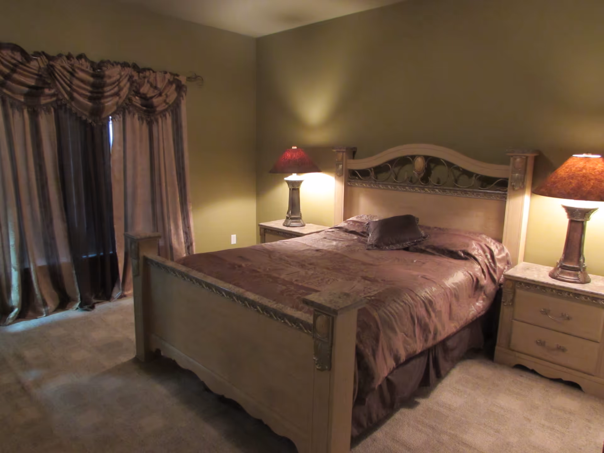 A warmly lit bedroom with a large wooden bed, matching nightstands and lamps, and heavy draped curtains.