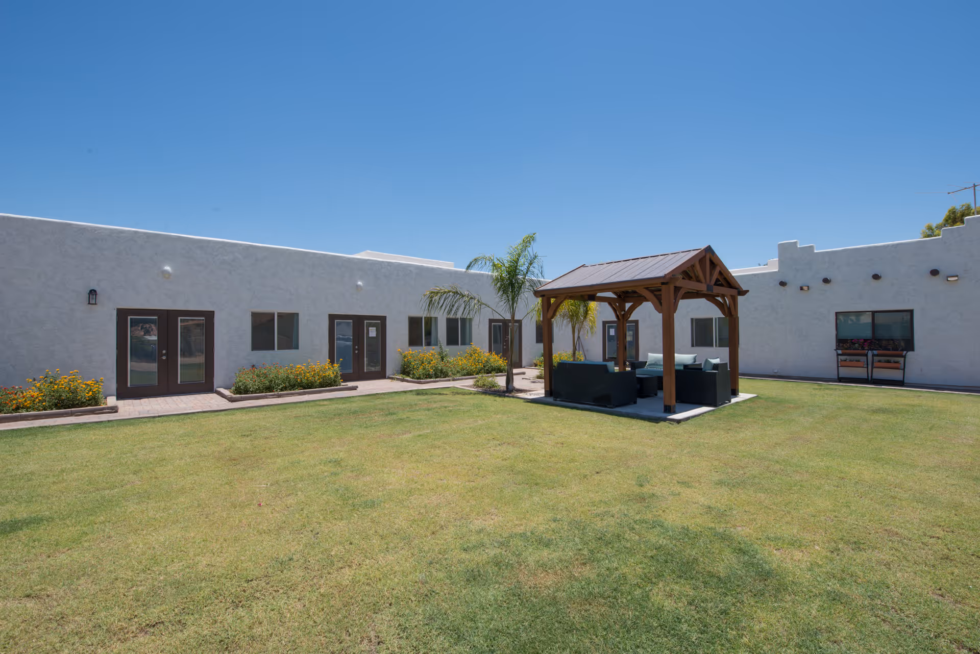 Outdoor courtyard area of Estancia Assisted Living with a green lawn, a wooden gazebo with seating underneath, surrounded by a single-story white building with multiple doors and windows, under a clear blue sky.