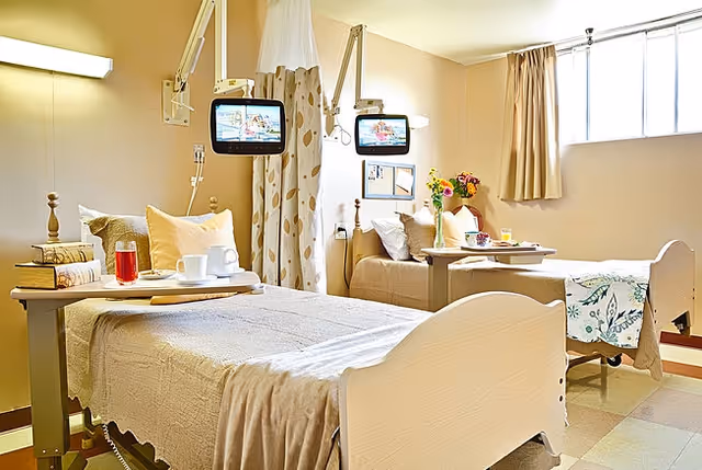 A bright and clean room with two hospital-style beds separated by a curtain. Each bed has a small overbed table with items like a glass of juice, a cup, and a book. Mounted televisions are positioned above each bed. The room has a window with beige curtains and a vase of flowers on a table near the far bed.