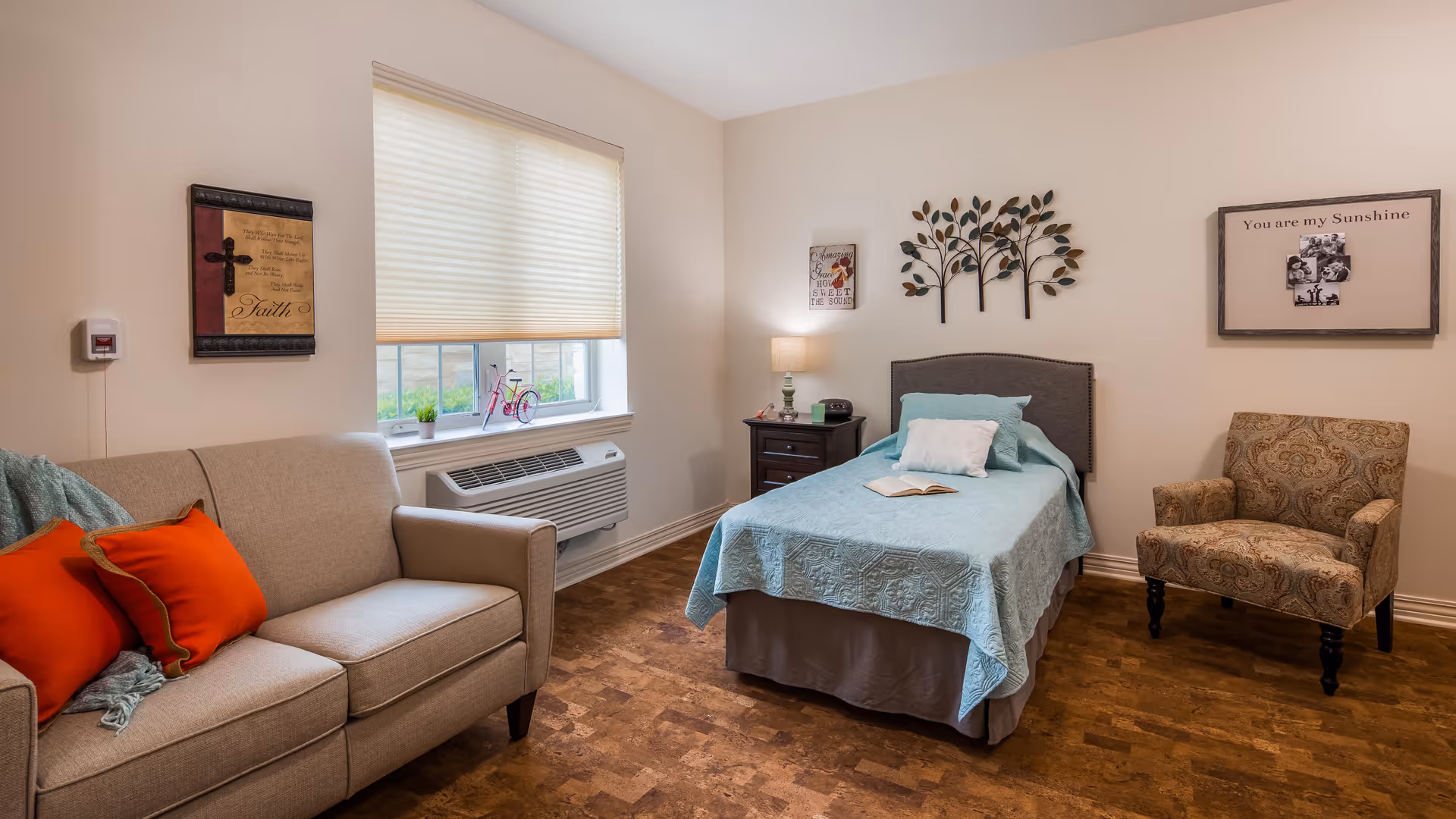 A cozy bedroom with a single bed covered in a light blue quilt, a beige sofa with orange pillows, a patterned armchair, a nightstand with a lamp and clock, wall decorations including a metal tree sculpture and framed pictures, and a window with a beige blind partially drawn.