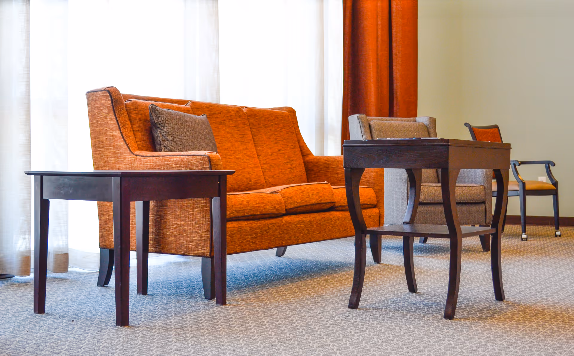 A cozy seating area with an orange upholstered sofa, a gray armchair, a wooden side table, and a wooden coffee table on a patterned carpet. Light filters through sheer white curtains with orange drapes on the side.