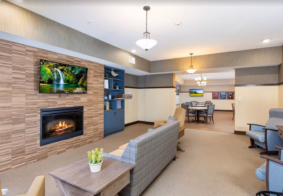 Cozy communal lounge with a fireplace, wall-mounted TV, upholstered seating and a dining area visible in the background.