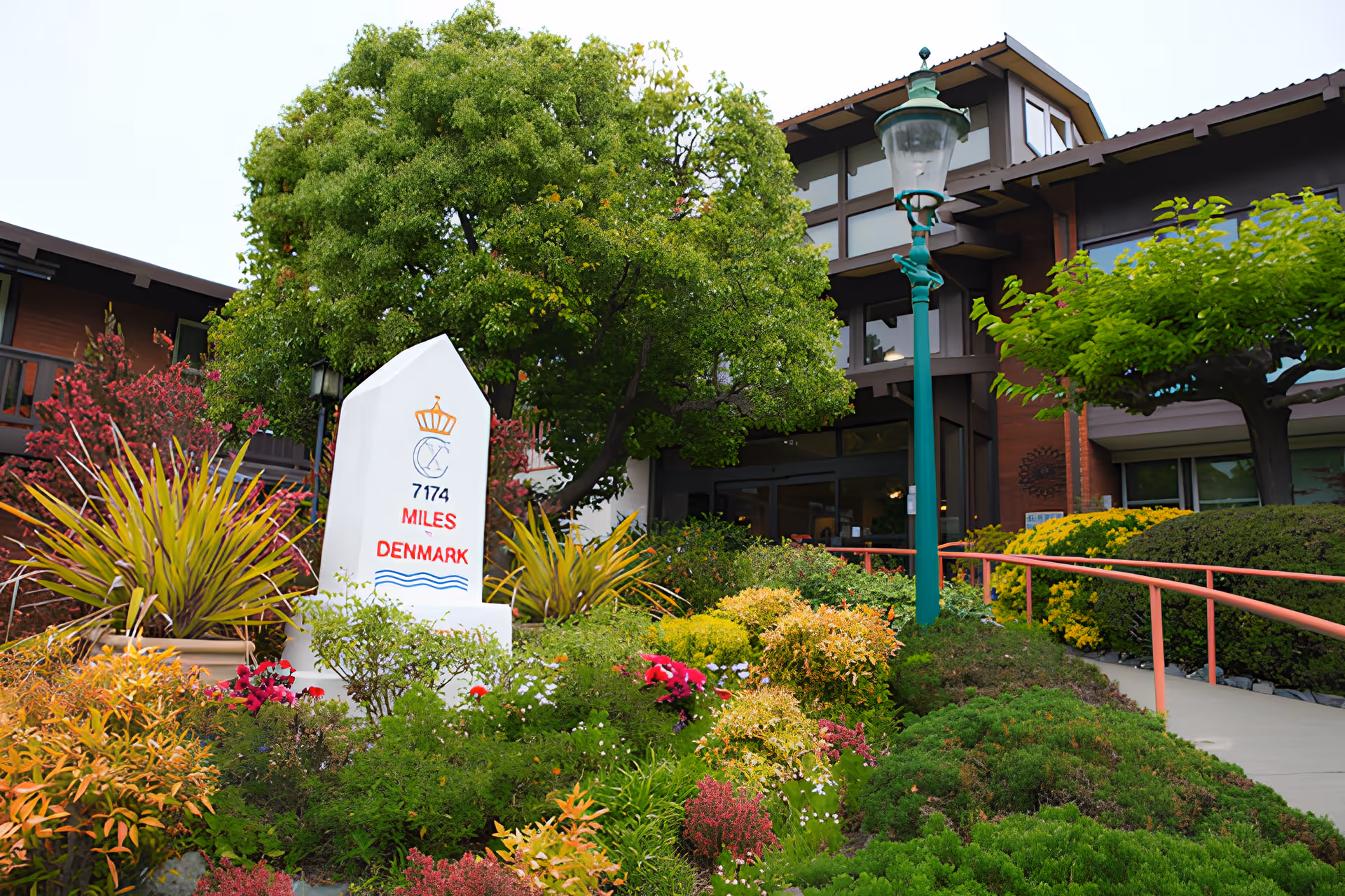 A landscaped garden area in front of a building with a variety of colorful plants and flowers, a white monument marker indicating 7174 miles to Denmark, a green lamppost, and a pathway with a red handrail leading to the entrance of the building.