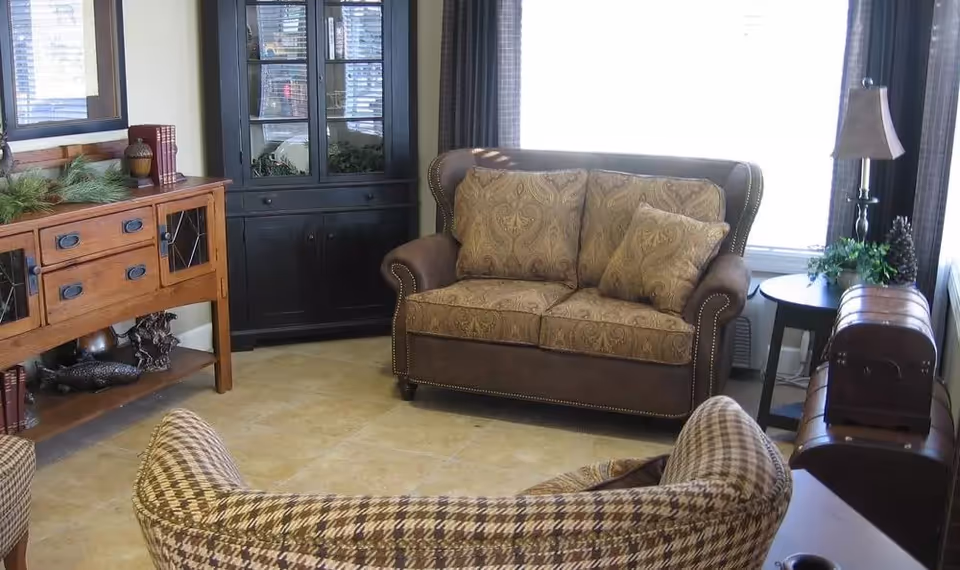A cozy living room with a patterned loveseat and matching armchair, a wooden sideboard with decorative items, a black cabinet with glass doors, a round side table with a lamp and plant, and a window with curtains letting in natural light.