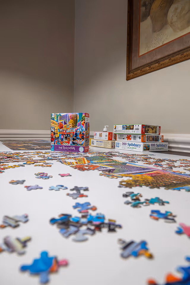 A partially completed jigsaw puzzle on a white surface with scattered puzzle pieces around it. Several puzzle boxes are stacked against a beige wall with a framed artwork hanging above.