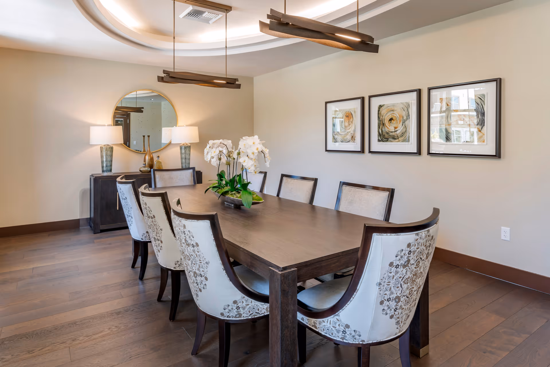 A modern dining room with a long wooden table surrounded by upholstered chairs, an orchid centerpiece, pendant lights, a round mirror and framed artwork.