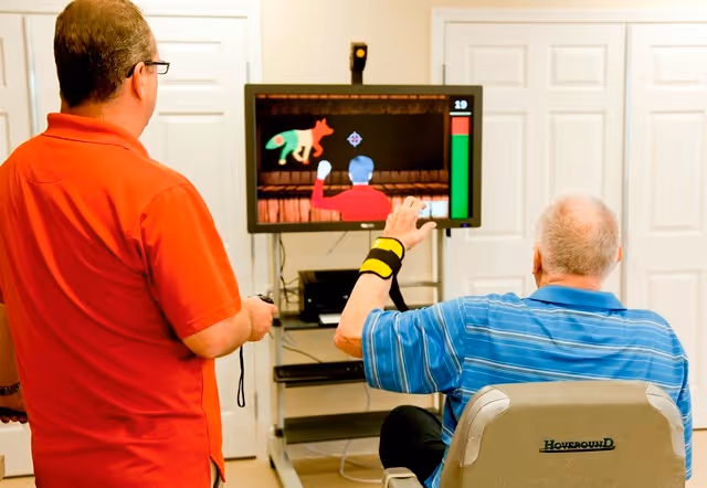Two elderly men playing a video game in a room with white paneled doors. One man in a red shirt is standing and holding a game controller, while the other man in a blue striped shirt is seated in a Hoveround mobility chair and wearing a wristband, interacting with the game on a screen mounted on a stand.