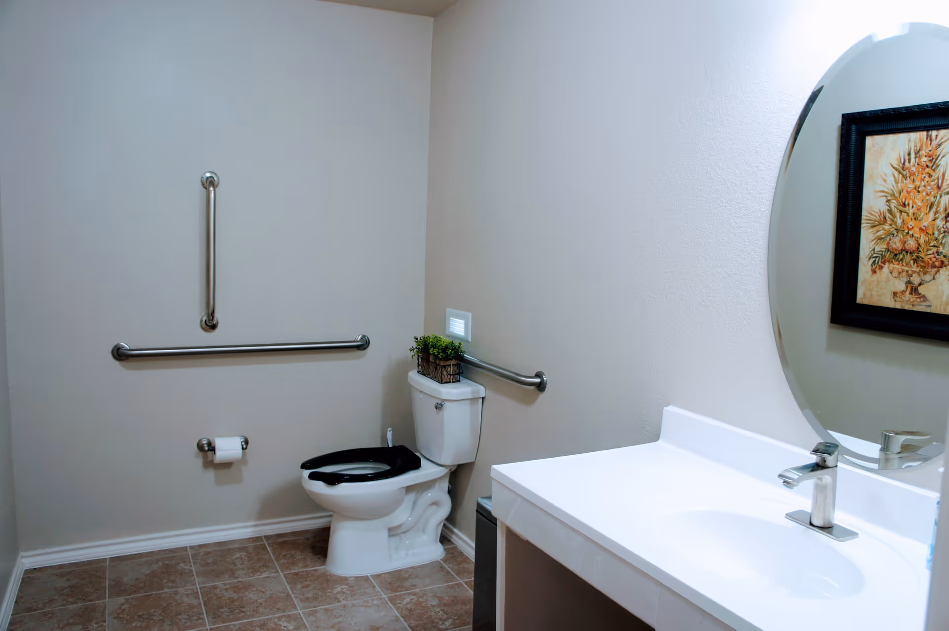 Accessible bathroom with a toilet (black seat), wall grab bars, a sink with faucet and round mirror, and a framed floral picture.