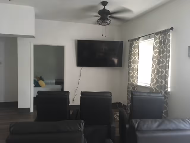 A living room area with four black leather chairs facing a wall-mounted flat screen TV. There is a ceiling fan above and a window with patterned curtains to the right. An open doorway on the left reveals a bedroom with a bed and pillows.