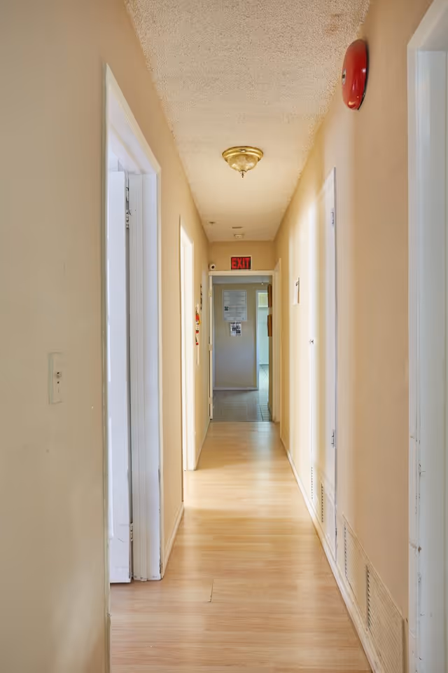 A narrow hallway with light beige walls and light wood flooring, featuring several white doorways on both sides. A red fire alarm is mounted on the right wall near the ceiling, and a ceiling light fixture is centered above. At the end of the hallway, there is an exit sign above a doorway leading to another room with tiled flooring.