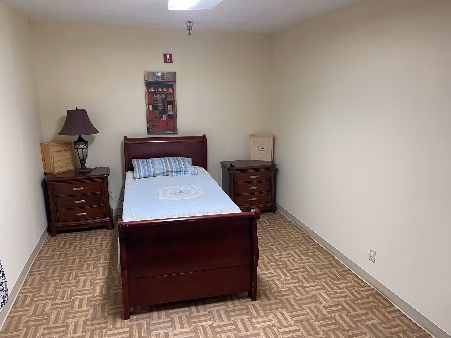 Small bedroom with a single wooden bed centered between two nightstands, a lamp, and wall art on a beige wall.