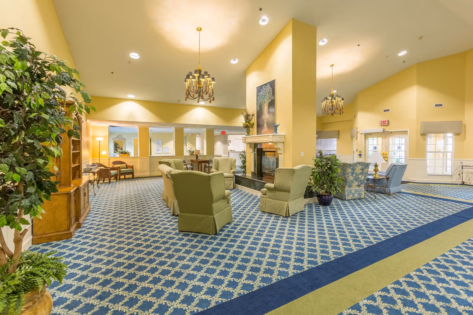 Bright lounge area with patterned carpet, multiple armchairs and sofas clustered around a central fireplace and chandeliers.