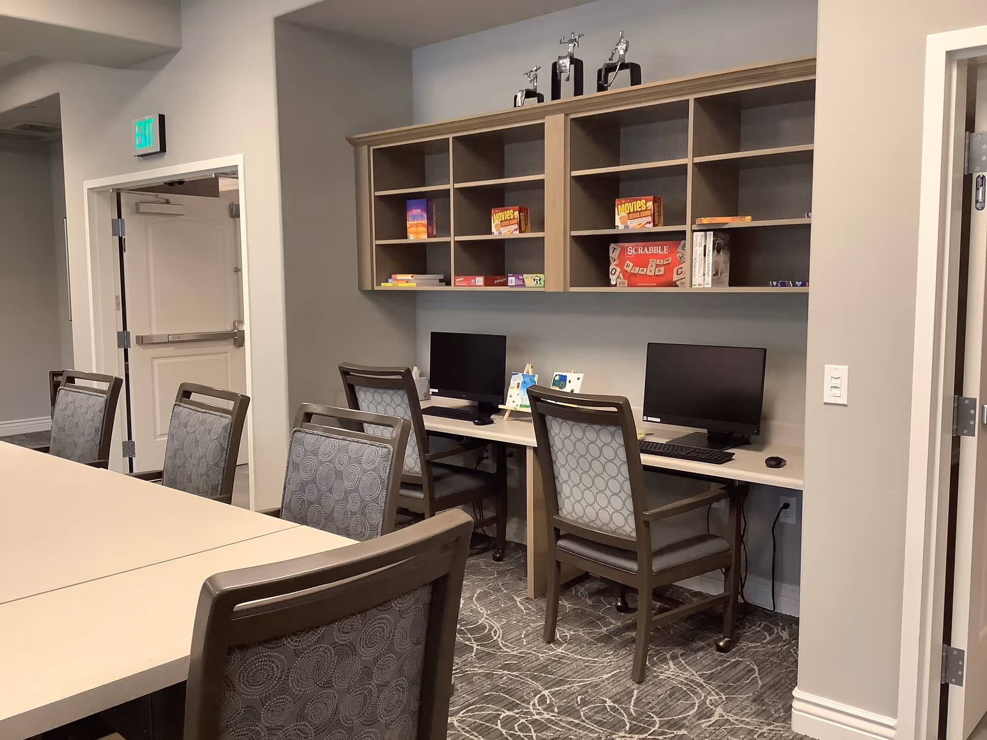 A room with a long table surrounded by chairs in the foreground and a desk area with two computer monitors and chairs against the wall. Above the desk are shelves holding board games and trophies. The room has neutral-colored walls and carpet with a swirling pattern.