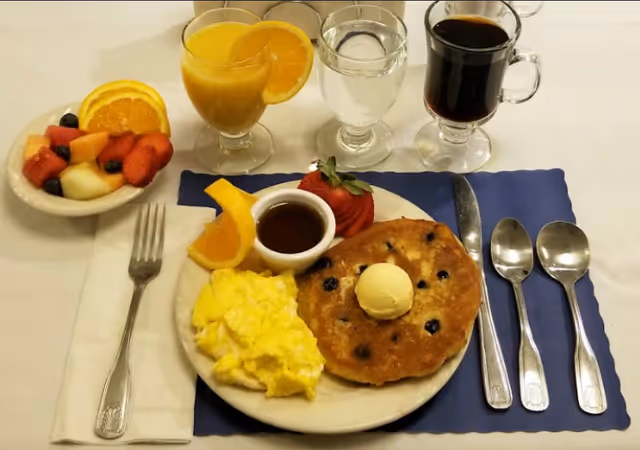 A breakfast plate with a blueberry pancake topped with butter, scrambled eggs, a small cup of syrup, fresh fruit, and glasses of orange juice, water, and coffee on a table.