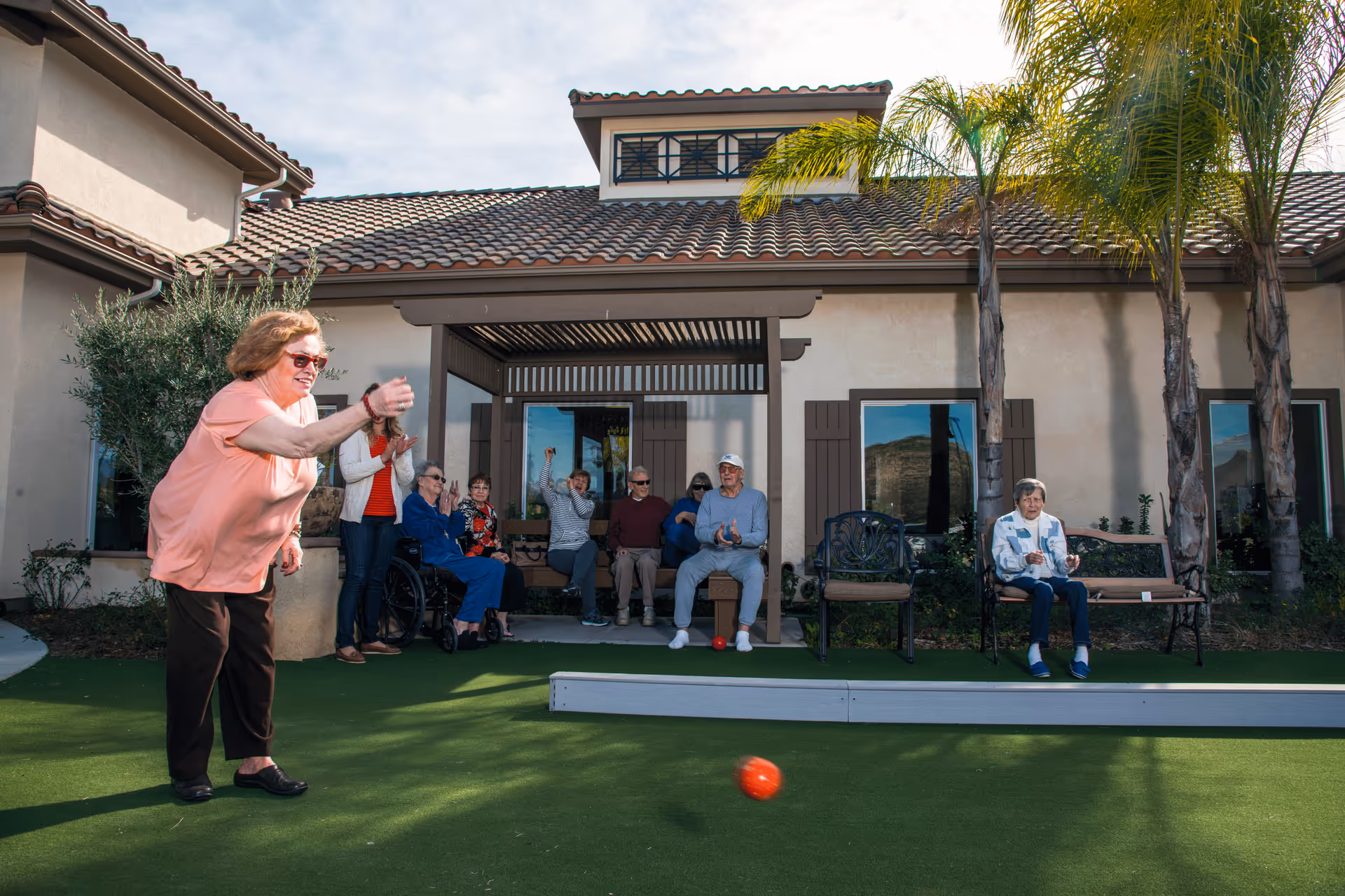 An elderly woman in a peach shirt and dark pants is playing bocce ball on a green lawn outside a building with a tiled roof. Several other elderly people are seated on benches and chairs under a shaded pergola, watching and cheering her on. Palm trees and shrubs are visible around the building.