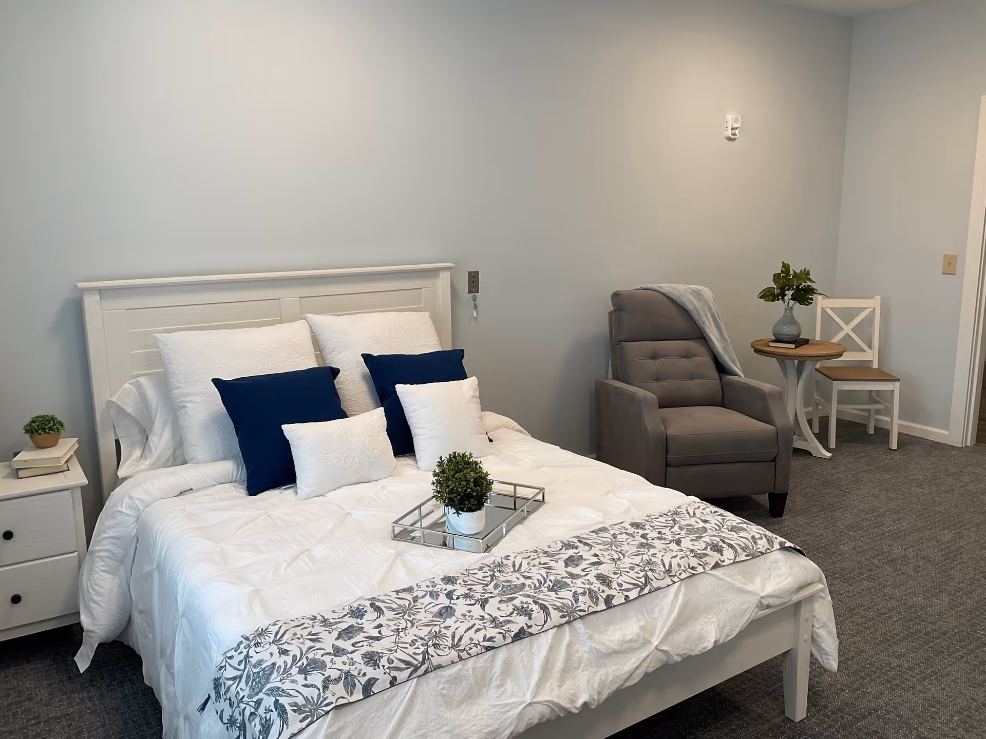 A neatly made bed with white and navy blue pillows and a white comforter with a floral patterned runner at the foot. A small potted plant on a mirrored tray sits on the bed. To the left of the bed is a white nightstand with two drawers and a small potted plant on top. To the right, there is a gray upholstered armchair with a light blue throw draped over the back, a round wooden side table with a vase of greenery and a book, and a white wooden chair. The room has light gray walls and gray carpeted flooring.