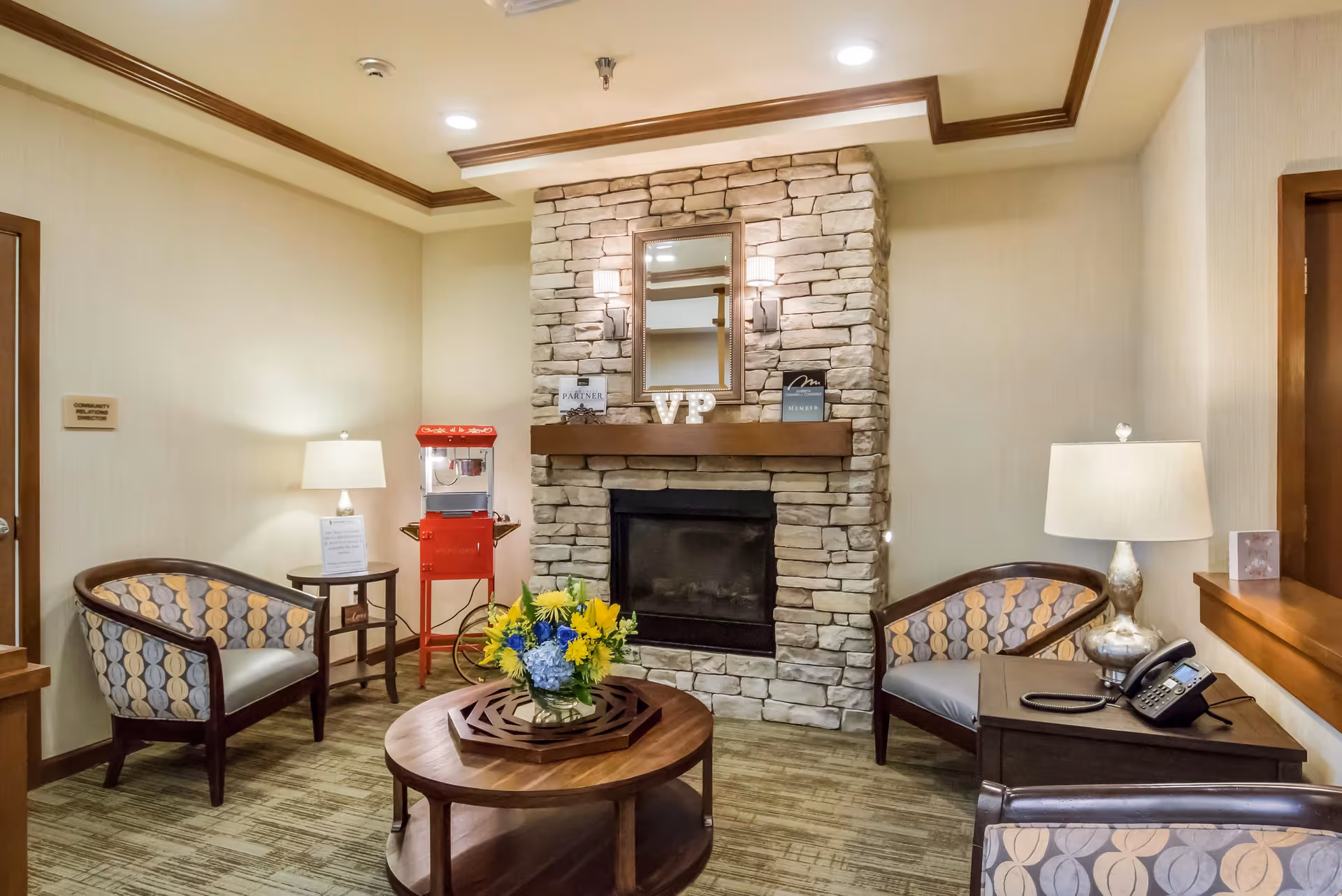A cozy sitting area with two patterned armchairs and a small table with a lamp on each side. A stone fireplace with a wooden mantle is centered on the wall, decorated with a mirror, small signs, and the letters 'VP'. A round wooden coffee table with a vase of yellow and blue flowers is in front of the fireplace. A red popcorn machine stands in the corner near a door labeled 'Community Relations Director'.