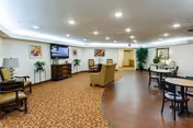 Well-lit senior living common area with seating, small dining tables, a television, and decorative plants.