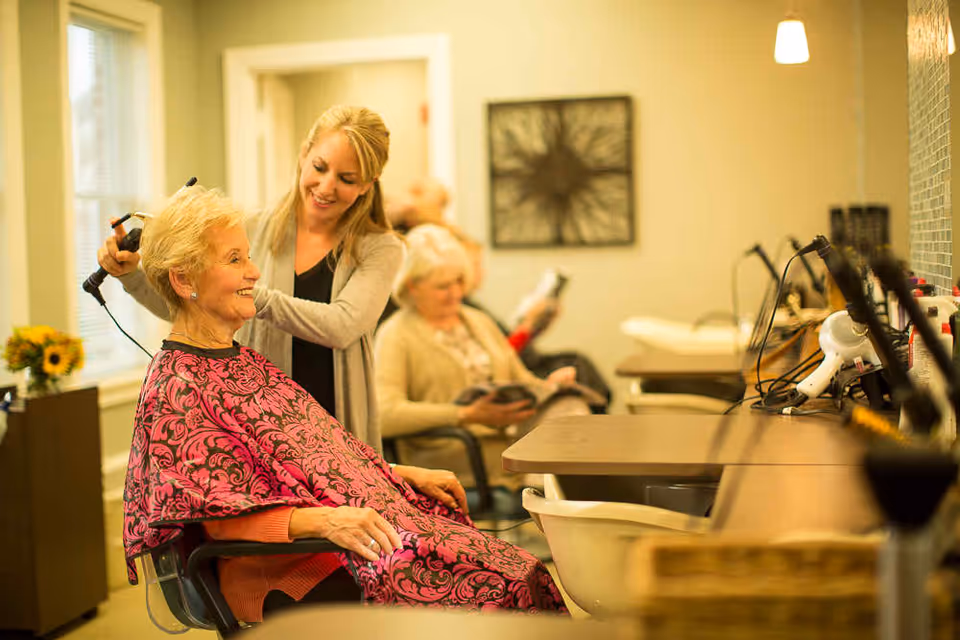 A hair stylist curling an elderly woman's hair in a bright salon area while another resident reads in the background.