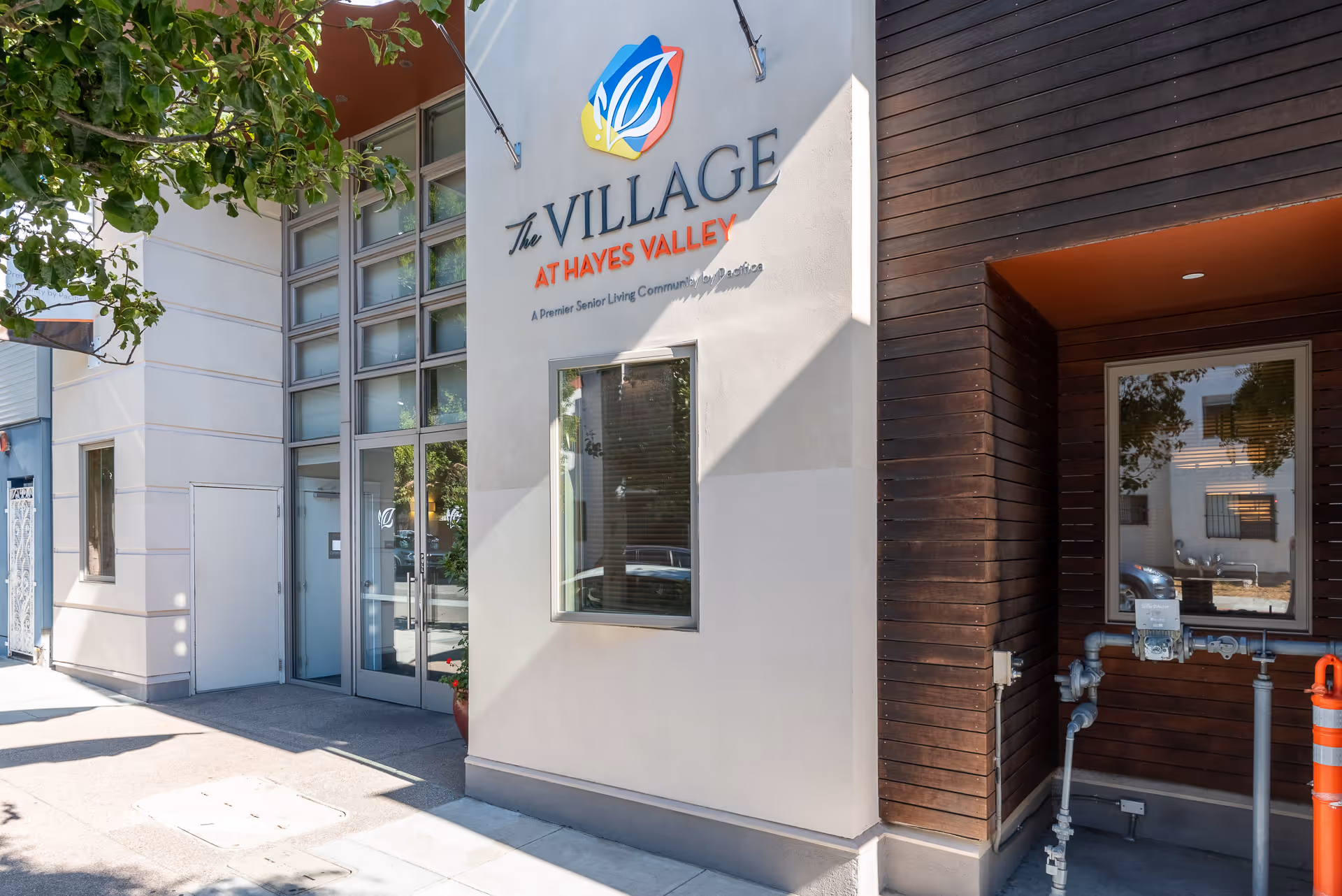 Exterior view of The Village at Hayes Valley senior living facility showing the entrance with glass doors, a window, and the facility's colorful logo and name on the wall. There is a tree casting shadows on the sidewalk in front.