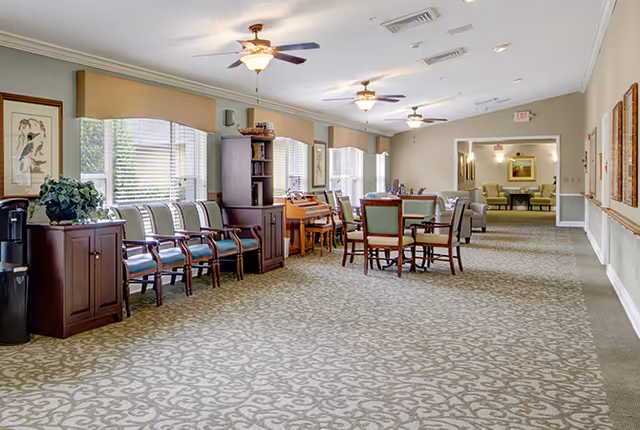 A spacious, well-lit common area in a senior living facility with patterned carpet, multiple ceiling fans with lights, and large windows with blinds. There are several wooden chairs with cushions lined up against the wall, a wooden cabinet with a plant on top, a piano, and a round table with chairs in the center. The far end of the room leads to another seating area with armchairs and framed artwork on the walls.