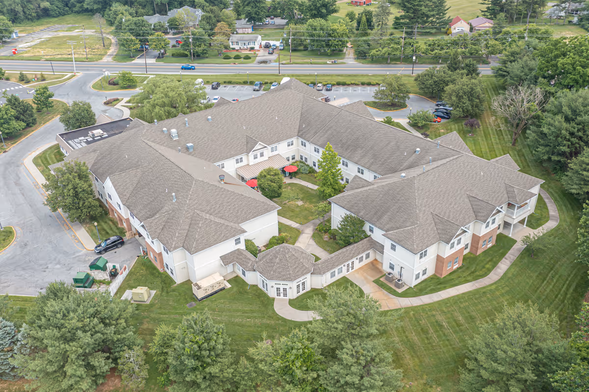 Aerial view of a U-shaped senior living facility building surrounded by lawns, trees, parking areas, and nearby roads.