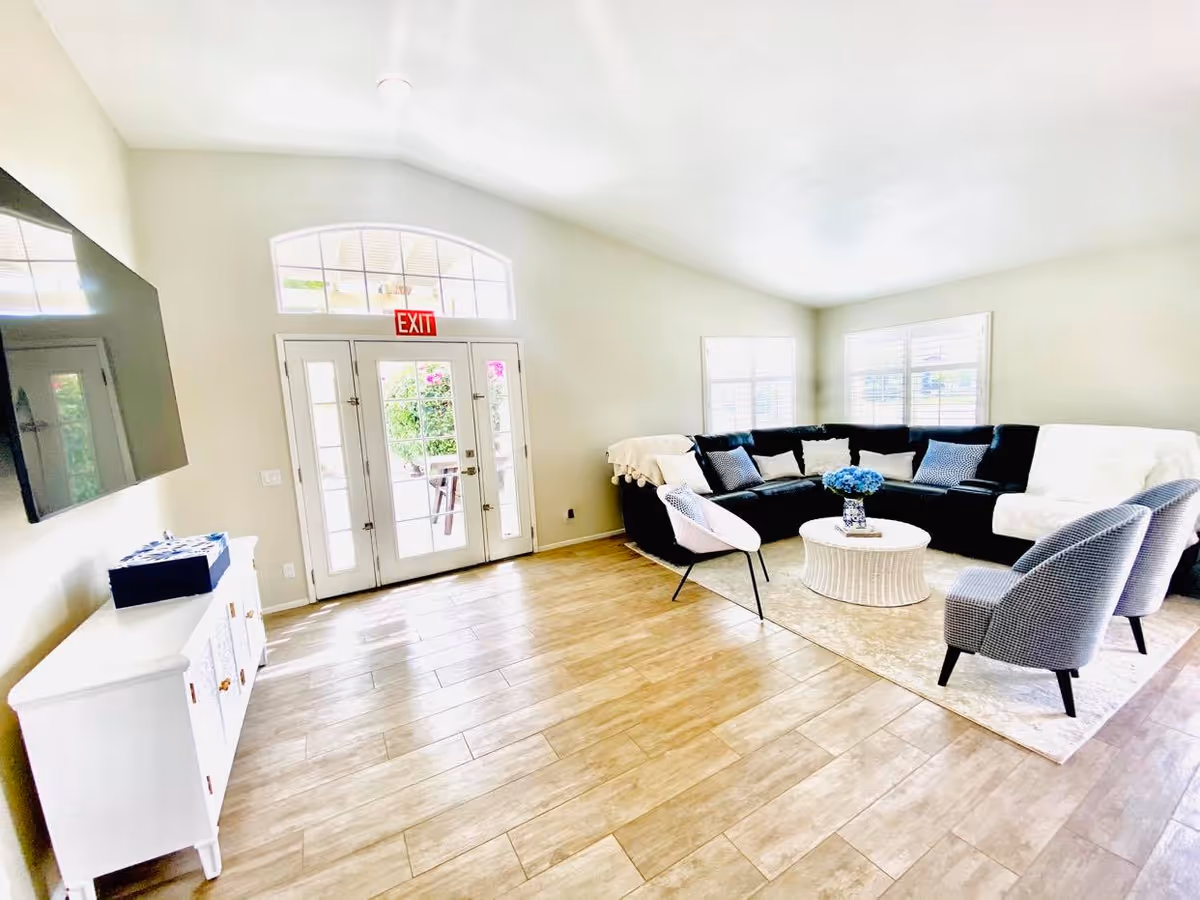 Bright living room with a large black sectional sofa adorned with pillows and white throws, two gray armchairs, and a white round coffee table with a blue flower arrangement. The room has light wood flooring, white walls, large windows with white blinds, a wall-mounted flat-screen TV, and a white cabinet. French doors with an exit sign above lead outside.