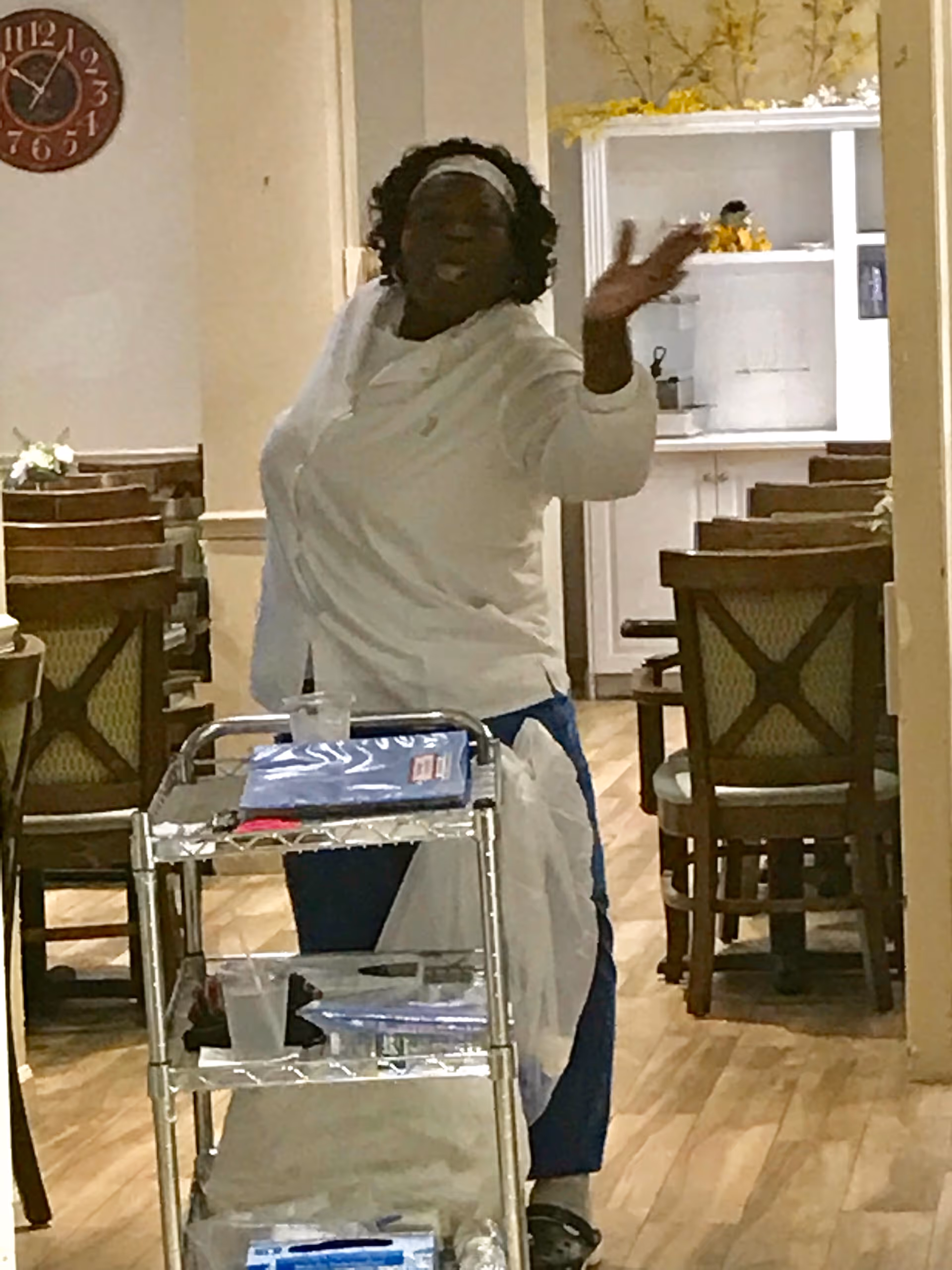 A woman wearing a white shirt and headband is standing behind a metal cart with various items on it in a dining area with wooden chairs and tables. The room has light-colored walls, a wooden floor, and a clock on the wall.