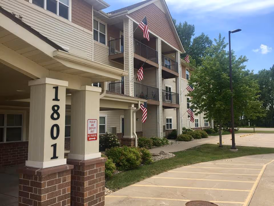 Exterior view of a multi-story senior living facility building with beige siding and brick accents. Several American flags are displayed on the balconies. The building number 1801 is prominently shown on a pillar near the entrance. There is a parking lot and a tree with green leaves near the building under a partly cloudy blue sky.