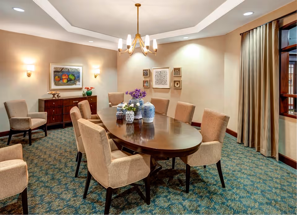 A well-lit dining room with a large oval wooden table surrounded by eight beige upholstered chairs. The table is decorated with several blue and white ceramic vases and a small flower arrangement. The room has patterned carpet flooring, beige walls with framed artwork, wall sconces, a chandelier, and a window with beige curtains.