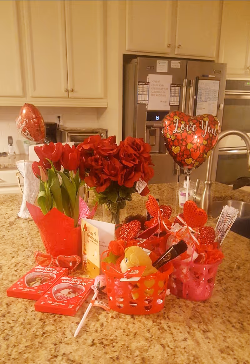 A kitchen countertop decorated with Valentine's Day items including red tulips, a bouquet of red roses, heart-shaped balloons with messages like 'Love You', Valentine's Day cards, red heart-shaped glasses, and small gift baskets filled with various items. The background shows a stainless steel refrigerator, oven, and cream-colored cabinets.