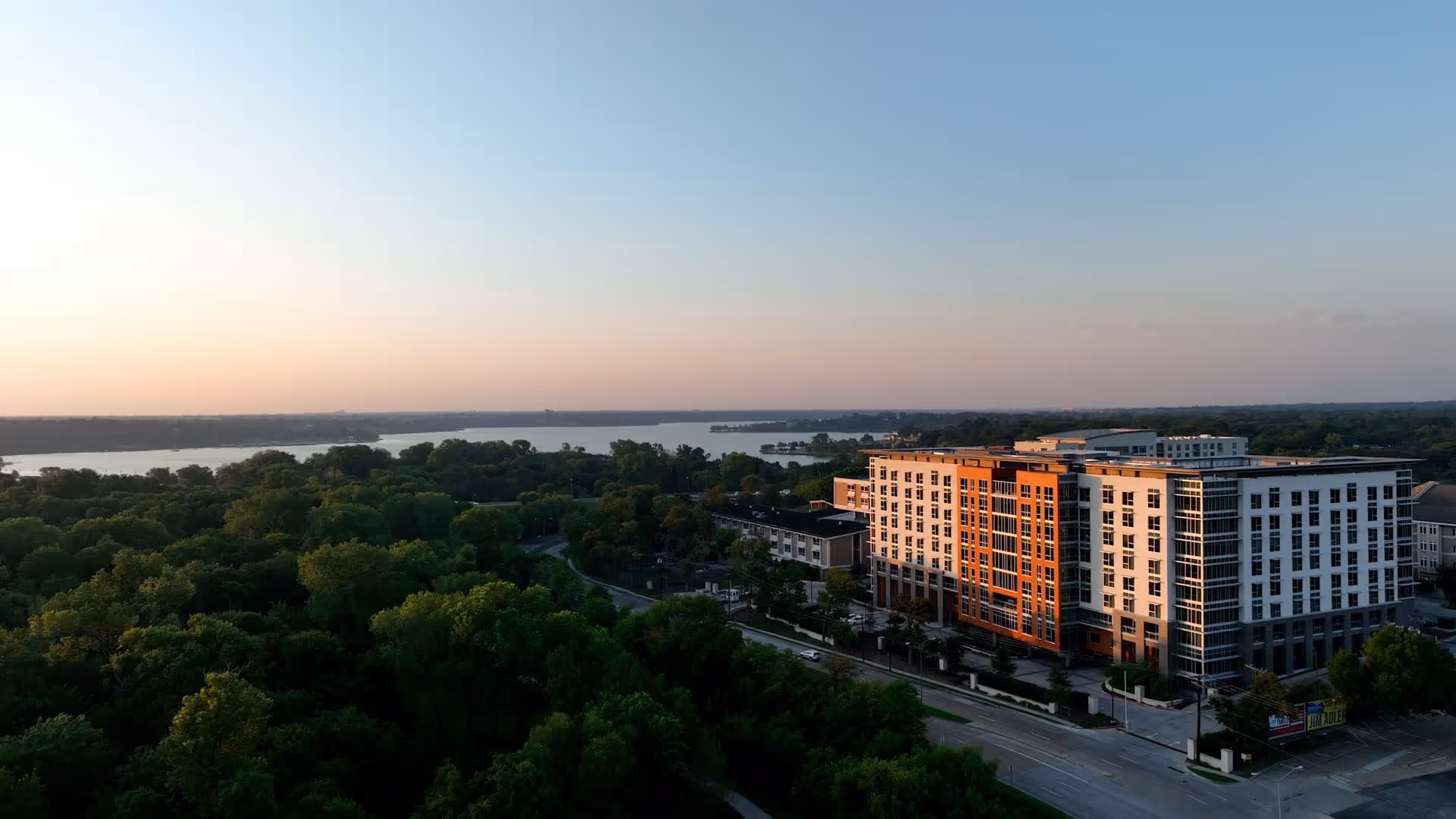 Aerial view of a large multi-story residential building next to a tree-lined park and a lake at sunset.