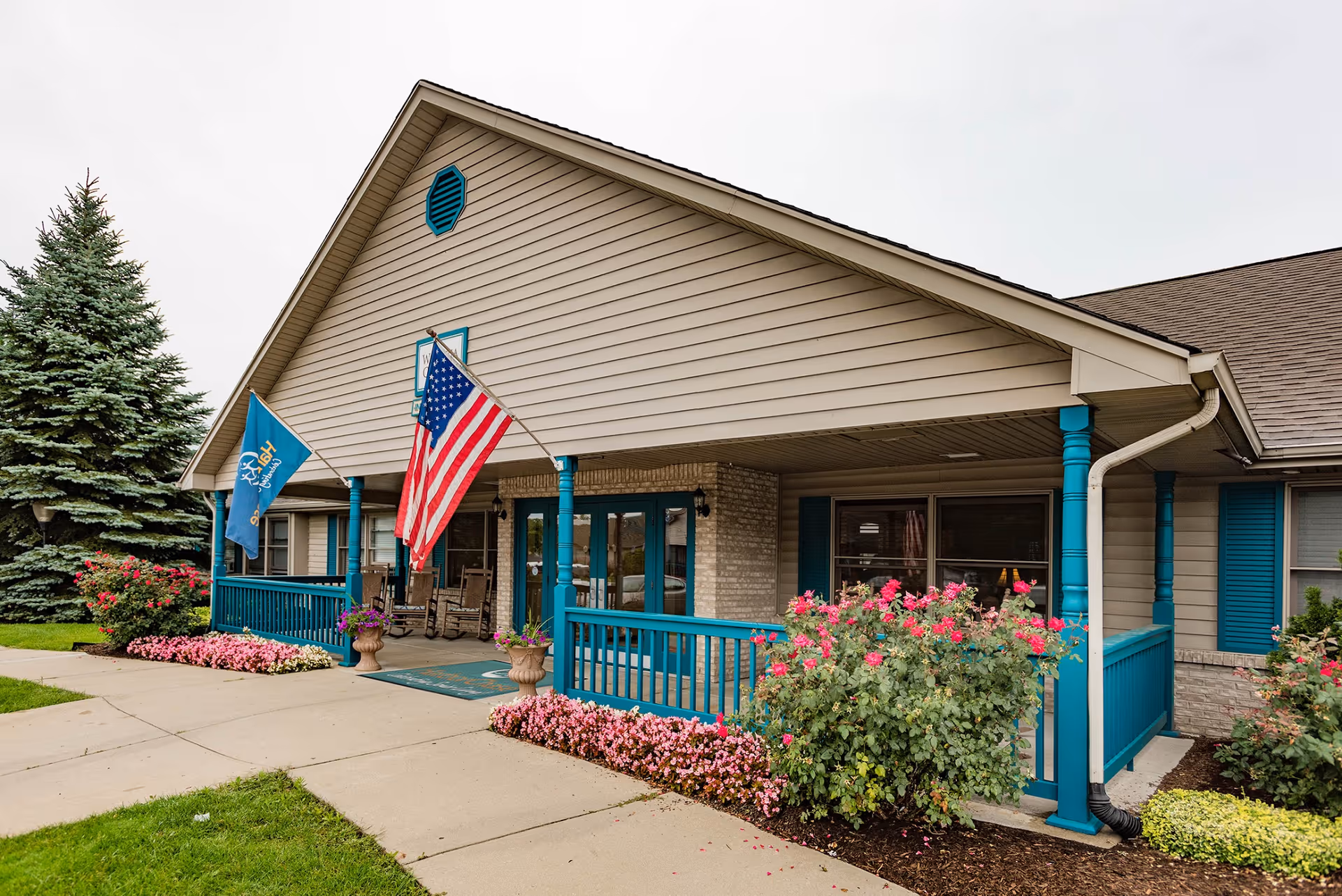 Exterior view of HarborChase of Sterling Heights building with a covered porch, two flags (American and HarborChase), rocking chairs, and landscaped flower beds with pink flowers and green shrubs.