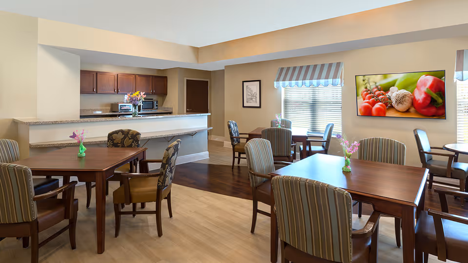 Bright common dining area with multiple wooden tables and upholstered chairs, a serving counter and kitchenette in the background.