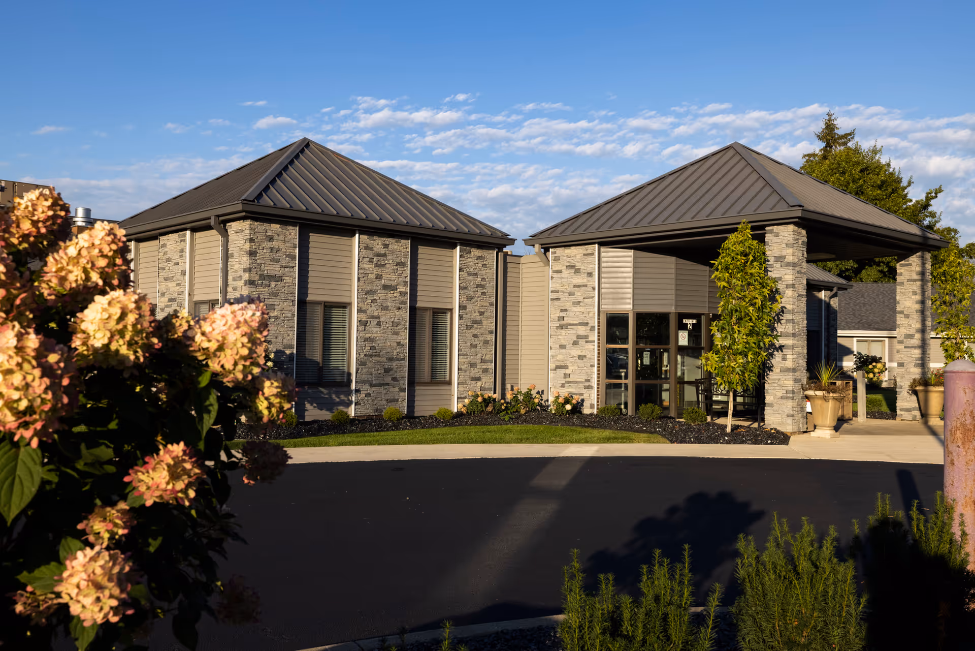 Exterior view of Caprice Senior Care Facility showing a modern building with stone and siding walls, metal roofs, a covered entrance, and landscaped surroundings with flowering plants and trees under a partly cloudy sky.