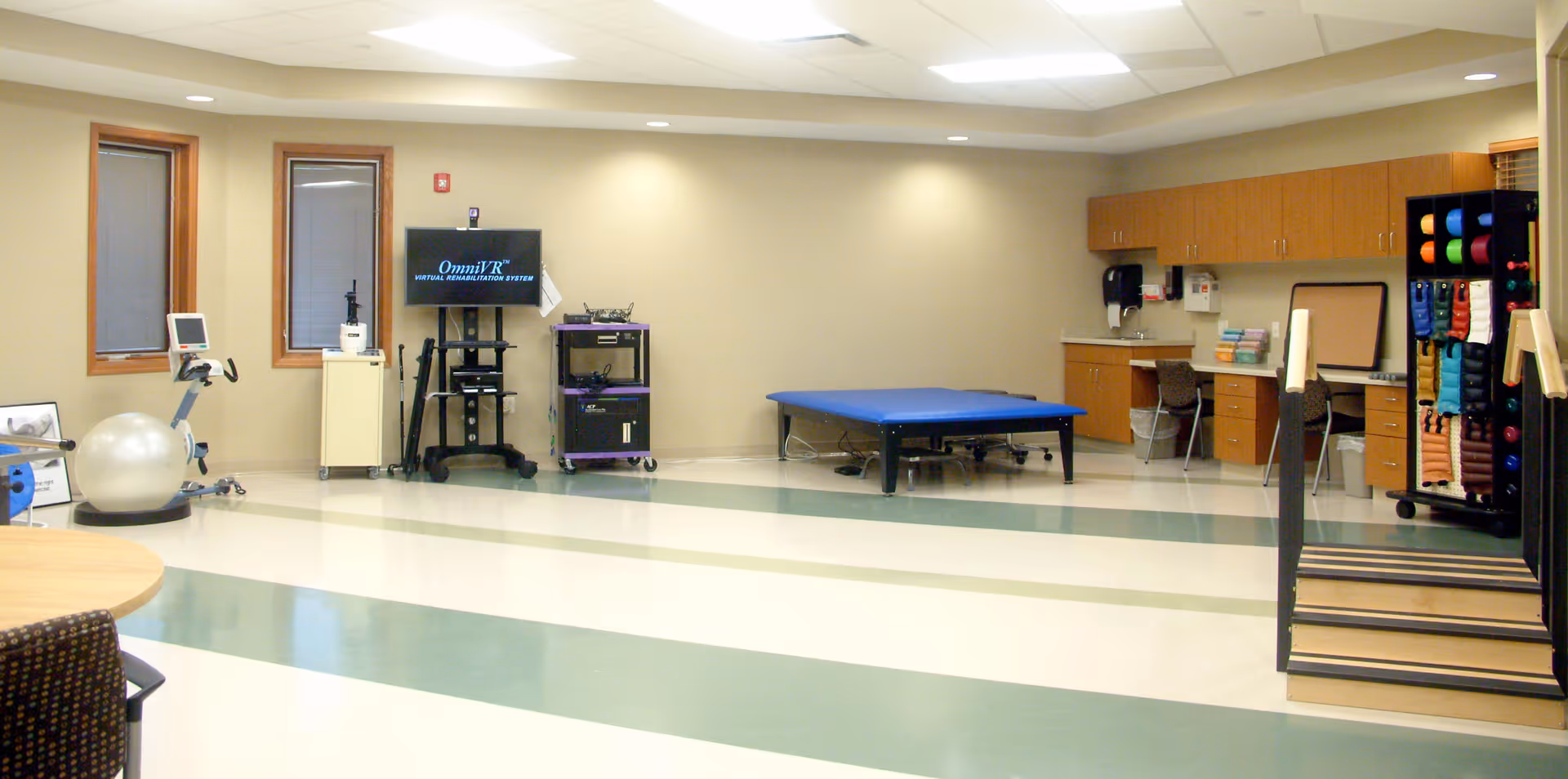 Spacious rehabilitation room with an exercise table, therapy equipment, stairs with handrails, exercise balls, and cabinets.