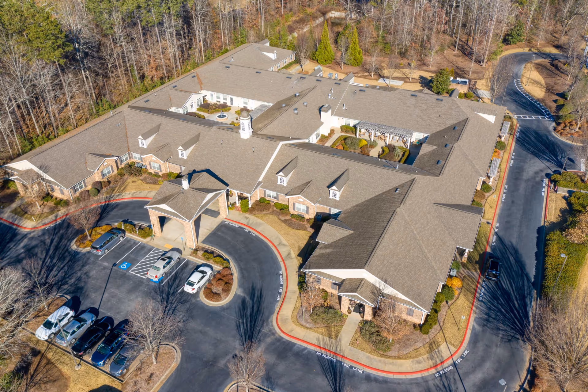Aerial view of Addington Place of Johns Creek senior living facility showing a large, single-story building with a covered entrance, surrounding parking lot with several cars, landscaped areas, and wooded surroundings.