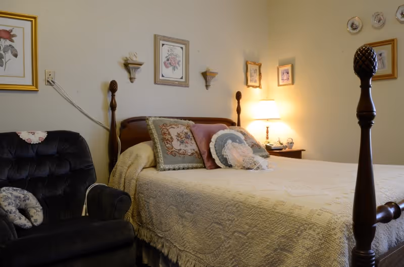 A cozy bedroom with a wooden four-poster bed covered with a cream-colored quilt and decorative pillows. To the left of the bed is a dark upholstered armchair with a small pillow. The walls are decorated with framed floral artwork and small shelves holding decorative items. A bedside table with a lamp and small decorative pieces is visible in the background.