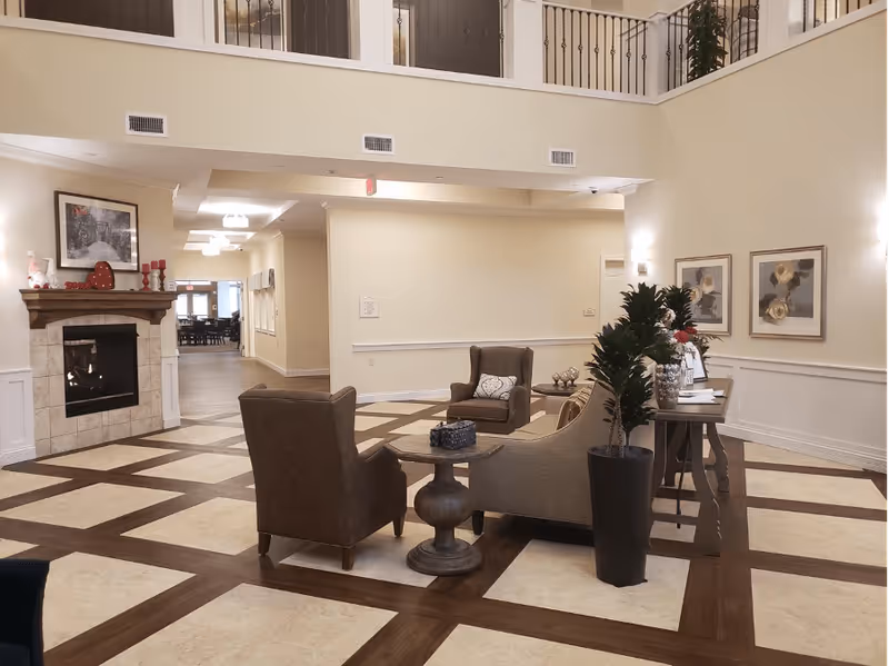 A spacious senior living facility common area with a high ceiling and balcony railing above. The room features a fireplace with decorative items on the mantel, two armchairs, a sofa, a small round table, a console table with flowers and decor, and framed artwork on the walls. The floor has a checkered pattern with wood and tile.