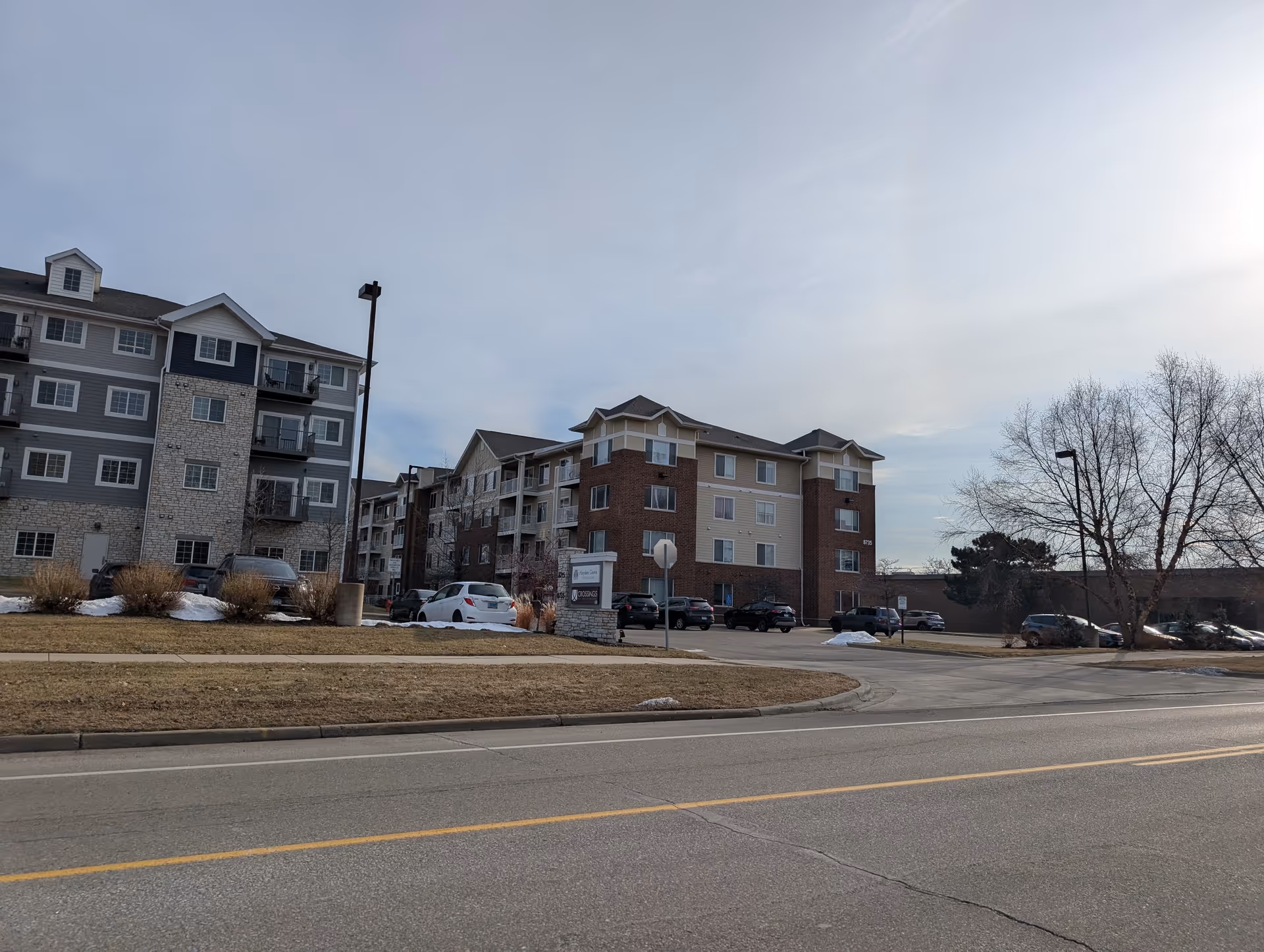 Front exterior view of a multi-story senior housing apartment building with parked cars and a road in the foreground.