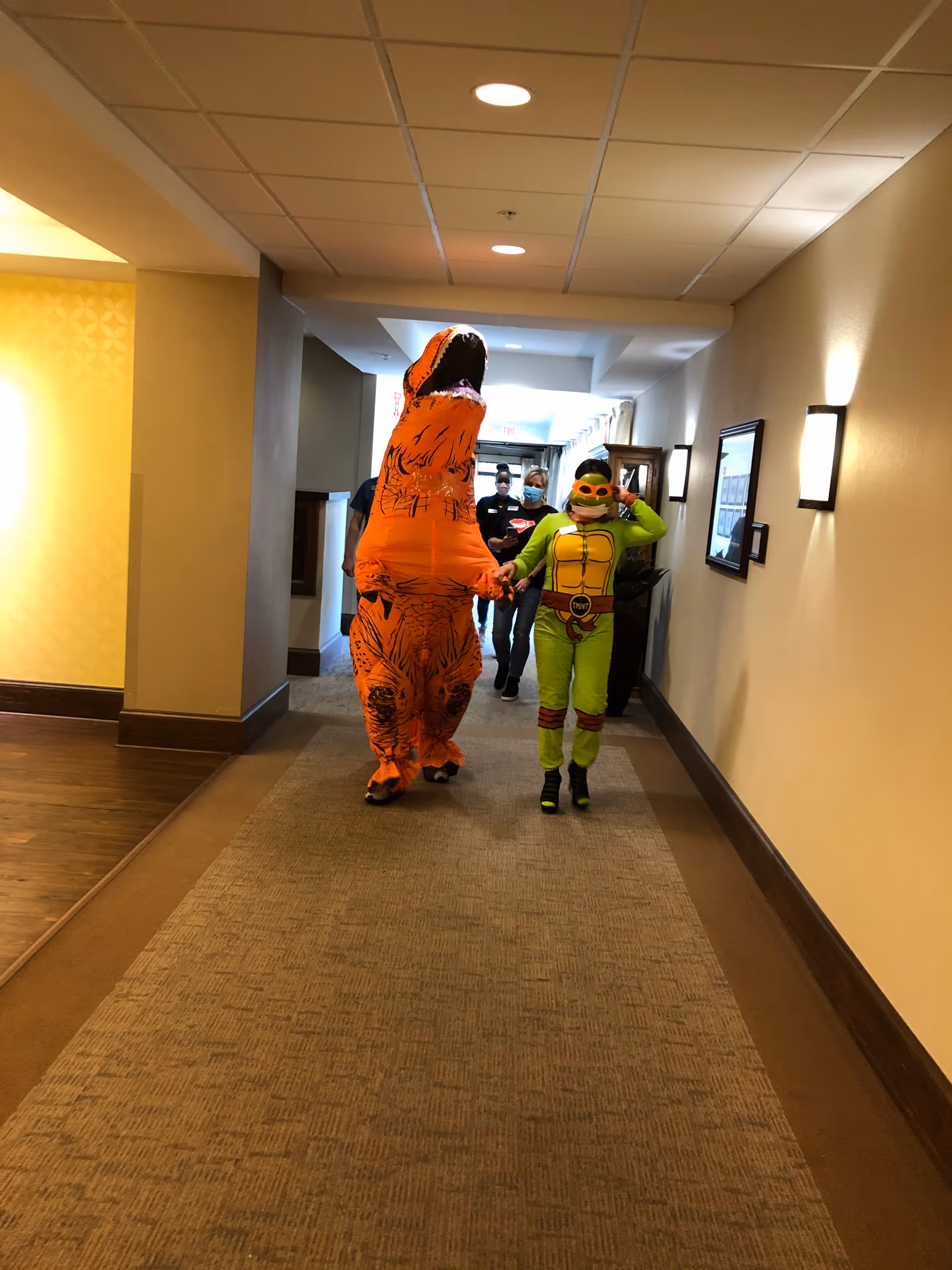 Two people walking down a hallway inside a senior living facility, one dressed in an inflatable orange dinosaur costume and the other in a green Teenage Mutant Ninja Turtle costume. Two staff members wearing masks follow behind them.