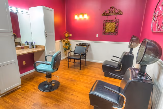 A bright pink salon room with styling chairs, hooded hair dryers, a vanity mirror and cabinets on a wood floor.