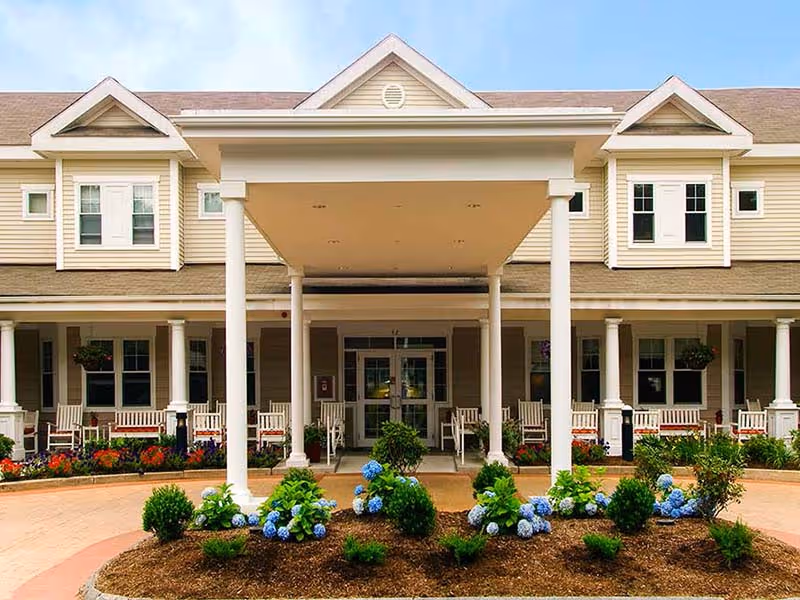 Front exterior view of a senior living facility with a covered entrance supported by white columns, beige siding, multiple windows, and a landscaped garden with green shrubs and blue flowers in front.