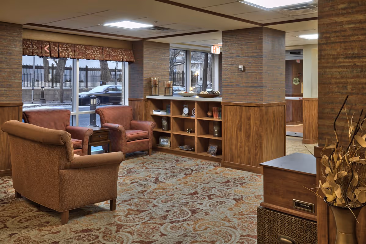 A cozy seating area in a senior living facility with three upholstered armchairs arranged around a small wooden side table. The space features wood-paneled walls, a patterned carpet, and a built-in wooden shelf with decorative items. Large windows provide a view of the outside street and trees.