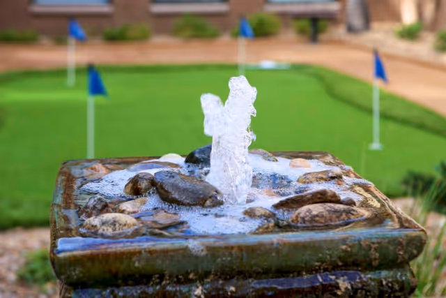 Close-up of a small water fountain with water bubbling up surrounded by rocks, with a putting green and several blue golf flags in the background.