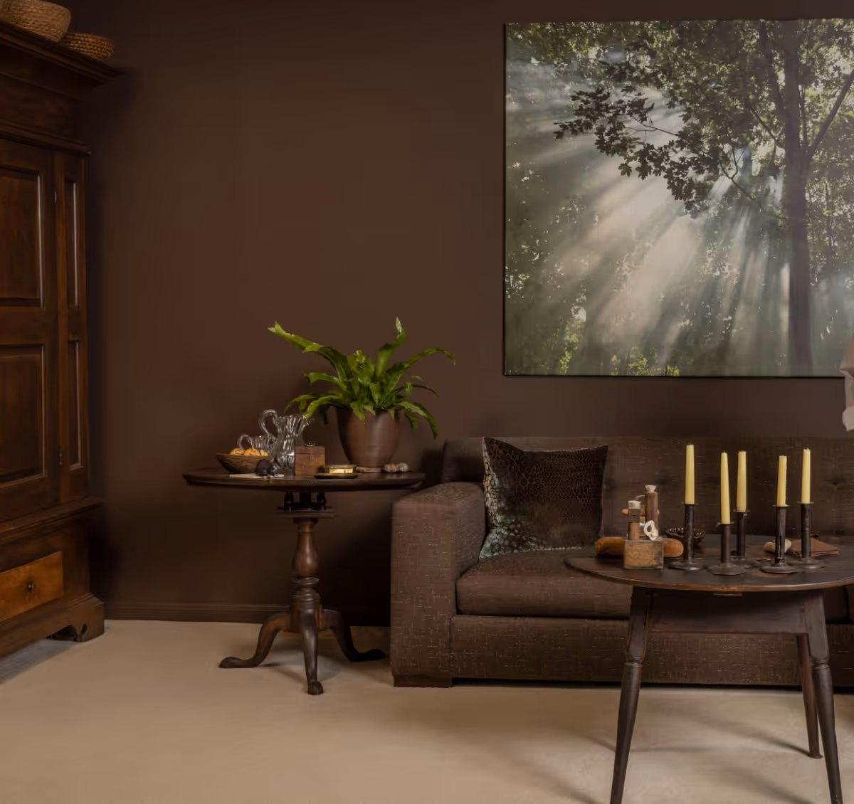 Cozy brown living room with a sofa, wooden side tables, candles, a potted plant, and large nature wall art.