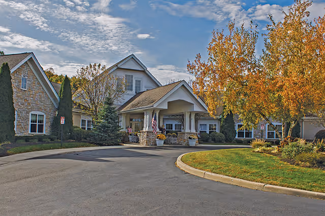 Exterior view of Buckingham Heights Memory Care facility showing a large building with stone and siding facade, a covered entrance with columns, an American flag, and landscaped grounds with trees displaying autumn foliage under a partly cloudy sky.