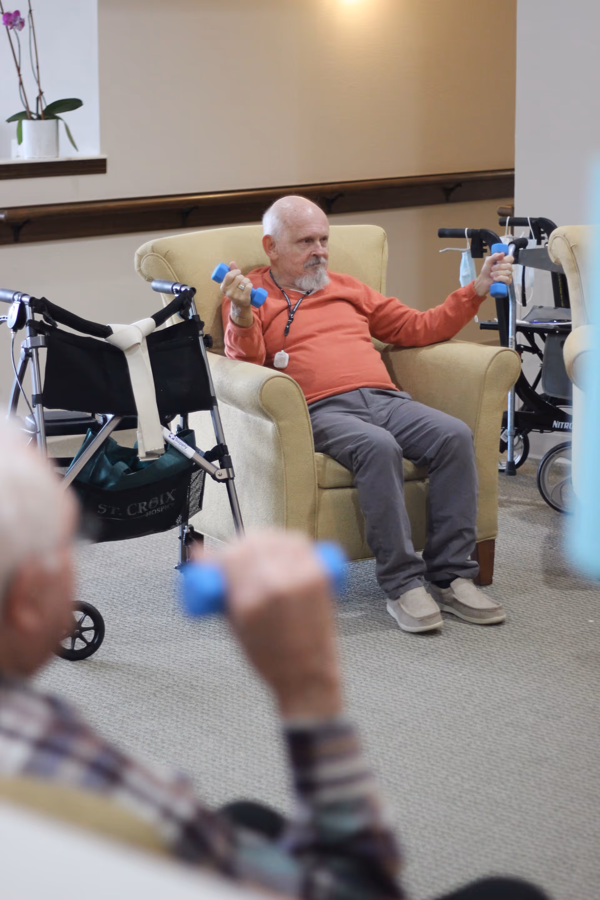 An elderly man in an orange long-sleeve shirt and gray pants sits in a beige armchair holding blue hand weights, exercising indoors. In the foreground, another person is partially visible also holding a blue hand weight. There are walkers and a beige armchair in the background, with a light-colored carpet and neutral walls.