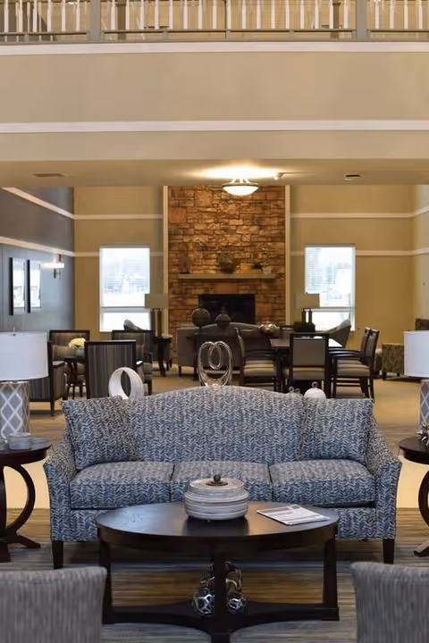 Communal living area with a patterned sofa and coffee table in the foreground, lamps and seating, and a dining area centered around a stone fireplace in the background.