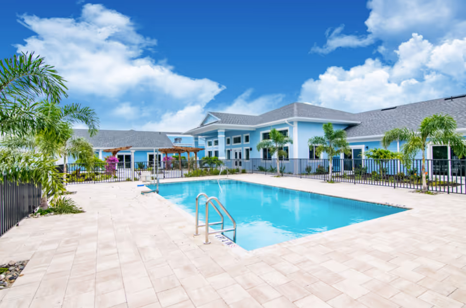 Outdoor swimming pool area at Hampton Manor of Cape Coral with a clear blue sky, surrounded by palm trees and a light blue building with a gray roof in the background.